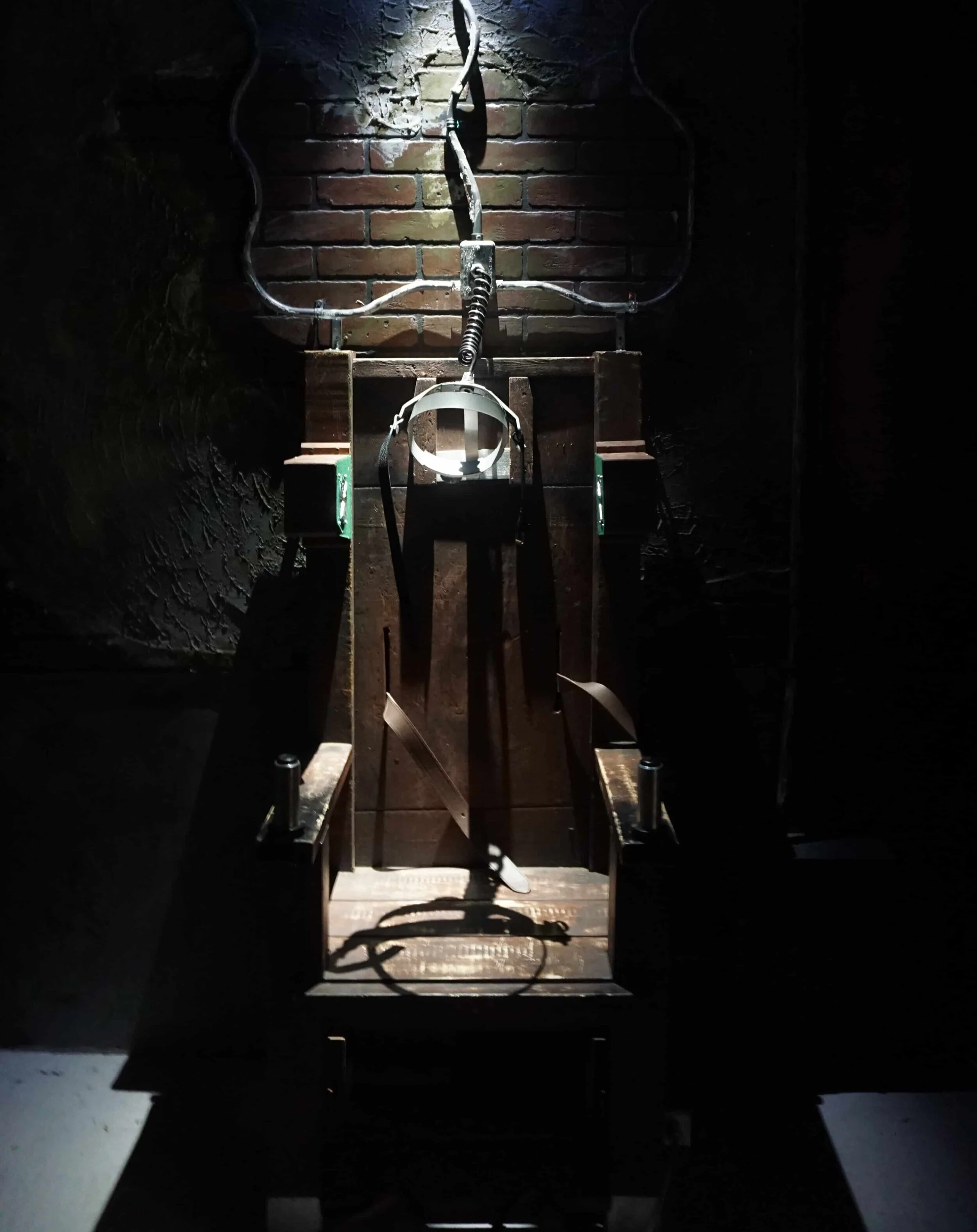 Wooden restraint chair with a metal headpiece and cables mounted against a brick wall.