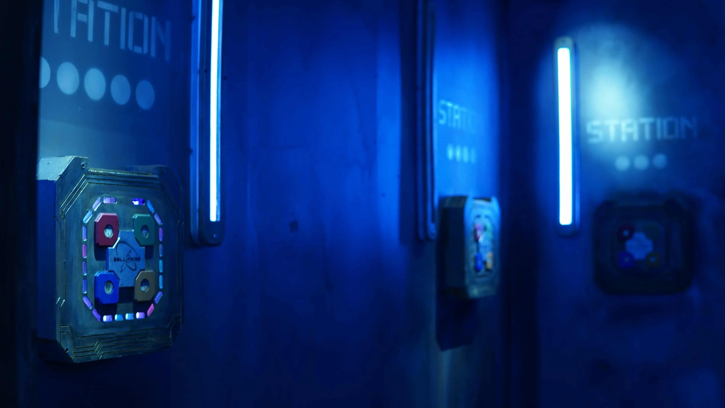 Blue-lit corridor with ‘STATION’ signage and illuminated wall-mounted puzzle boxes.