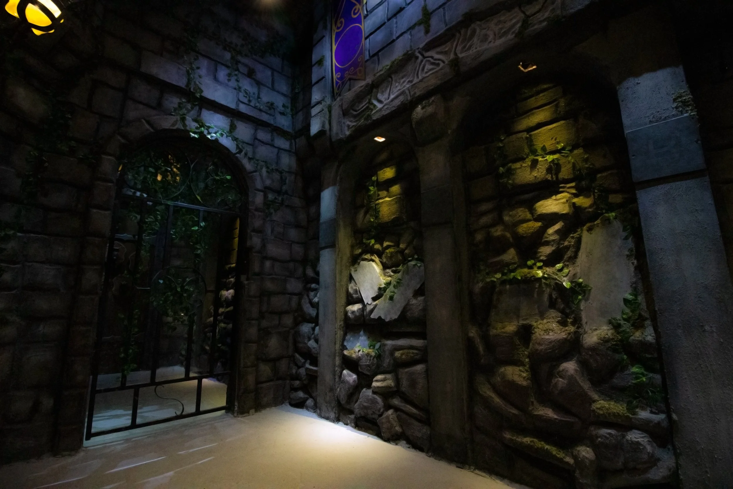 Moss-covered stone castle corridor with an arched doorway and iron gate lit by warm sconces.