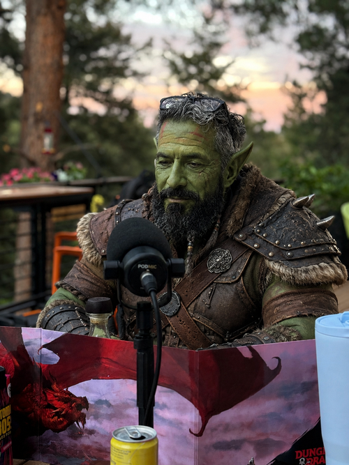 Markus Maximus, the Dungeon Master, drawn as a green-skinned orc in studded leather behind a microphone with a stack of rulebooks