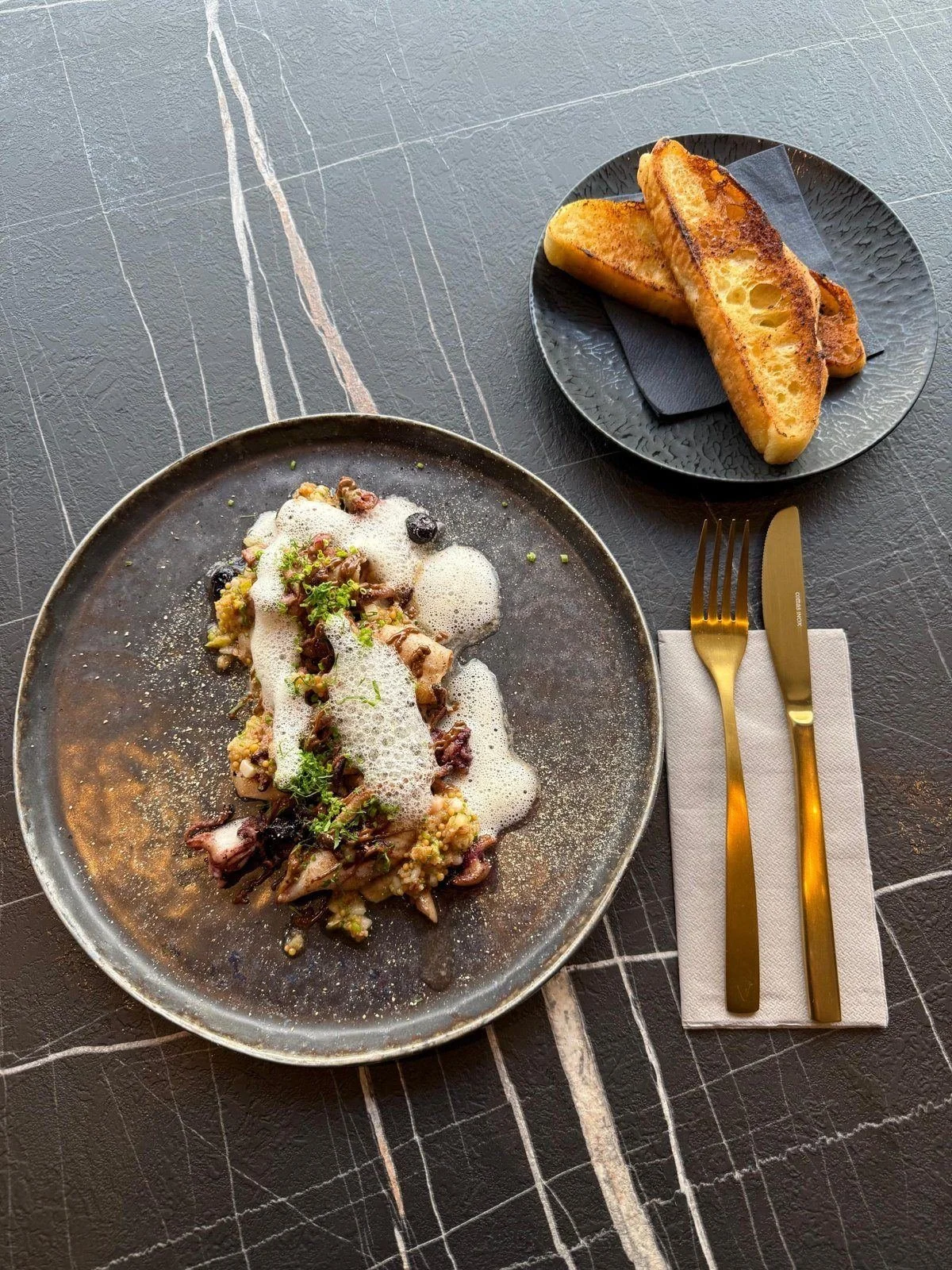 Tintenfisch mit Schaum und geröstetem Brot auf dem Marmortisch im Bistro, goldenes Besteck