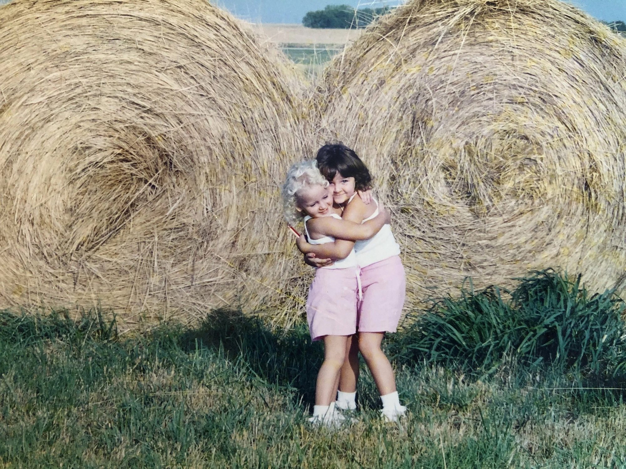Emma Grace and Lyla Belle as young girls, hugging in front of hay bales