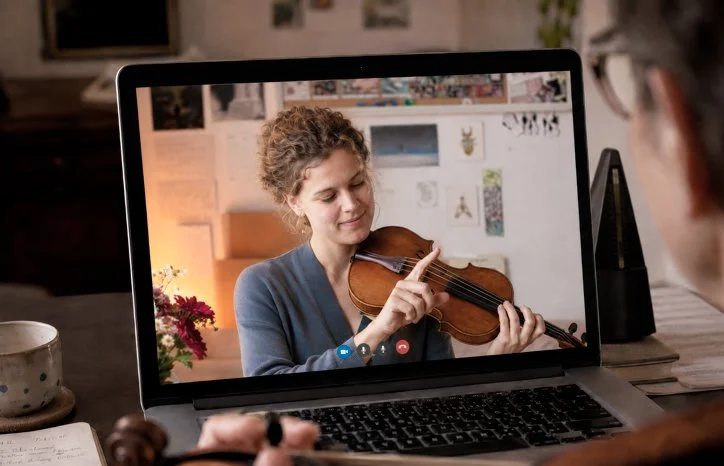 An online violin lesson on a laptop video call, the teacher pointing to a fingering position on the violin while an adult student watches from their desk.