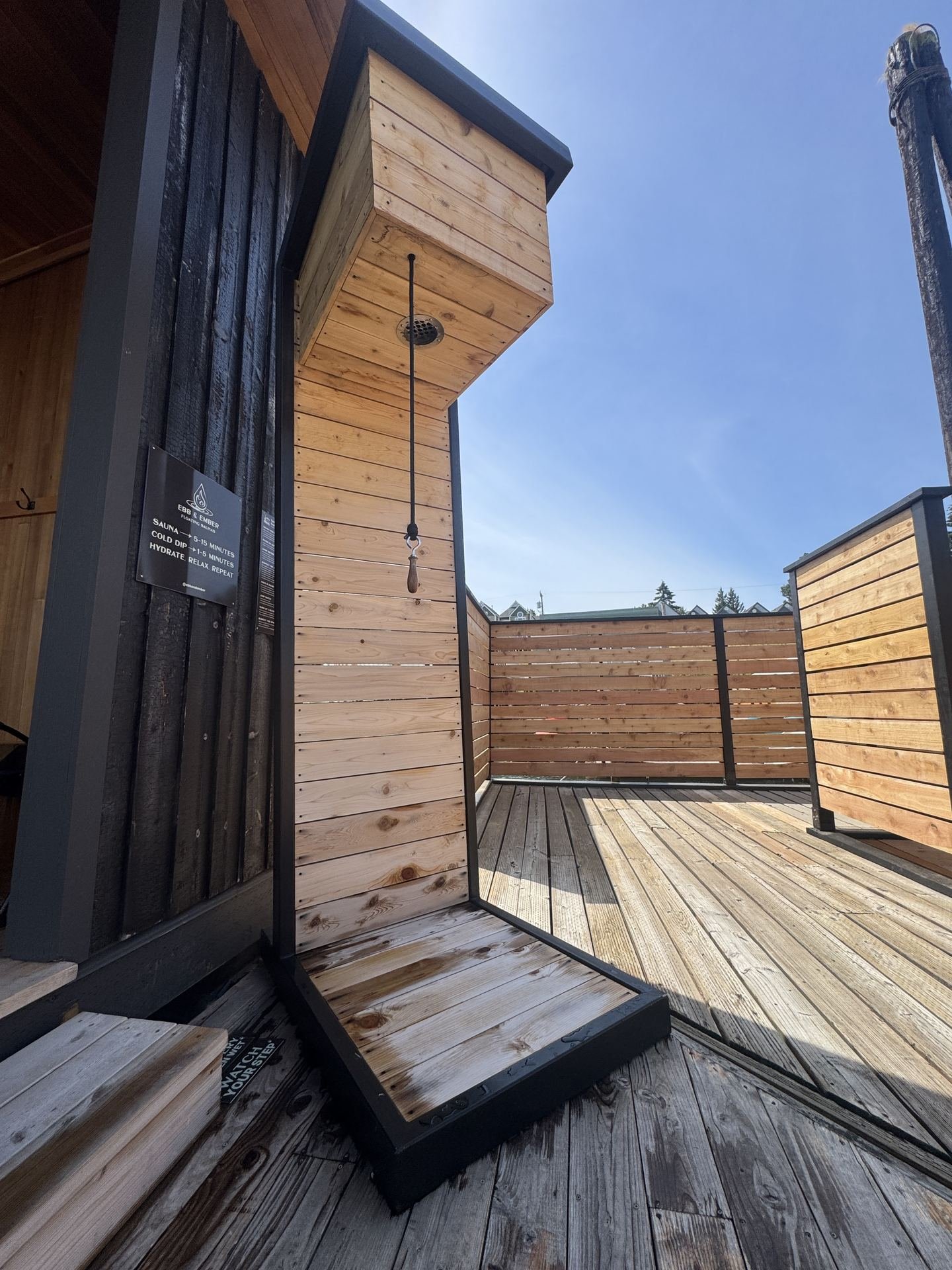 Cedar cold-rinse shower area at Ebb & Ember floating sauna against a blue sky