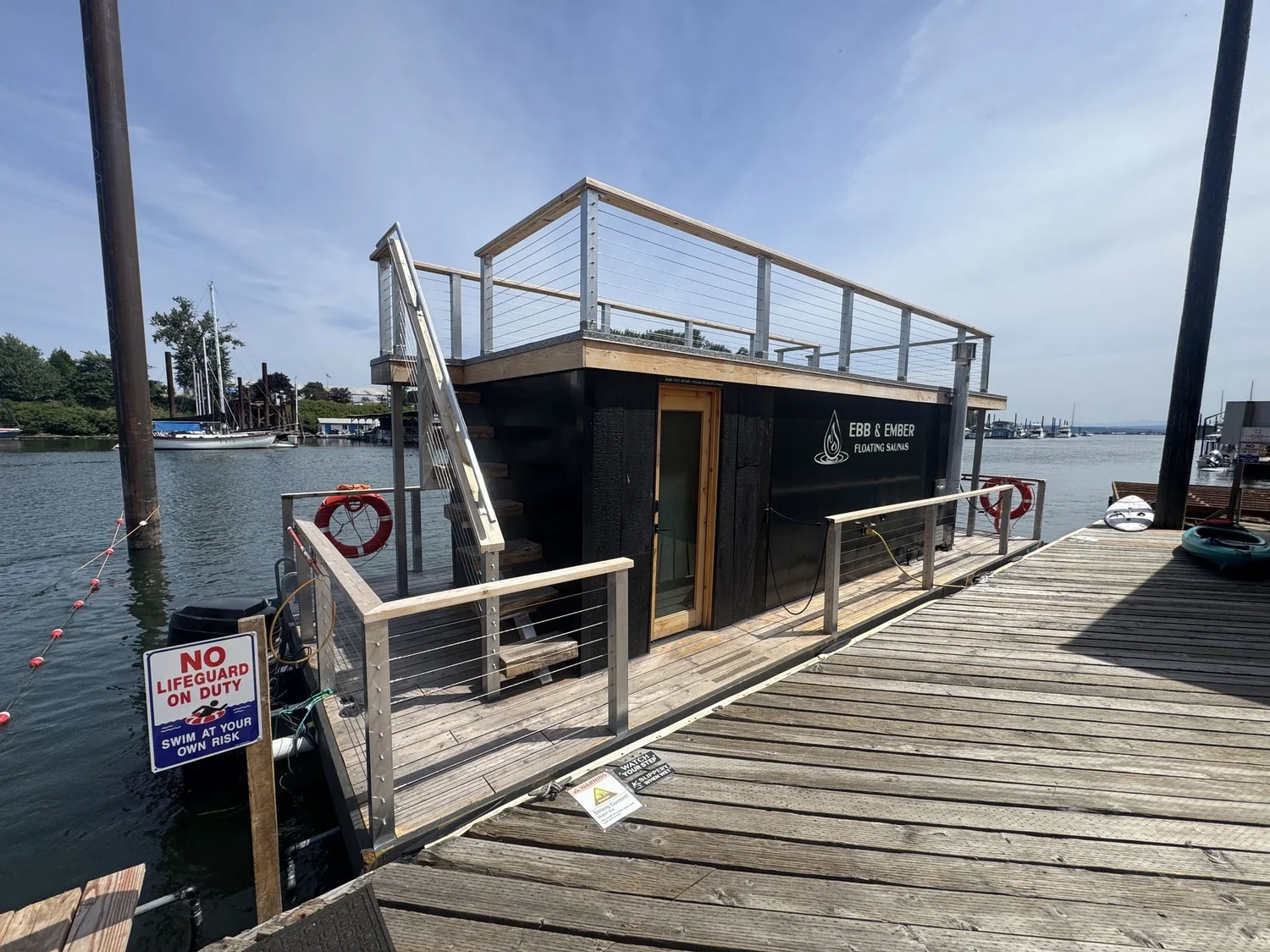 Exterior of the Ebb & Ember black floating sauna on a dock under a clear blue sky