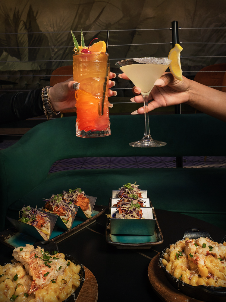 Two cocktails clinking over a table of tacos and mac and cheese