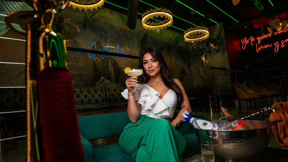 Guest on a green velvet sofa raising a cocktail under gold chandeliers
