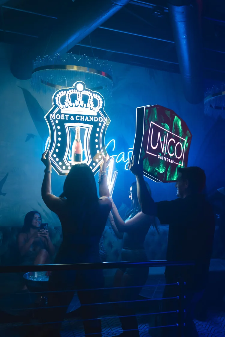 Guests carrying a lit Moët & Chandon sign and a Unico sign through the crowd under blue light