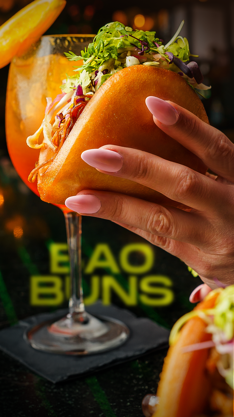 Hand holding a bao bun in front of an orange cocktail