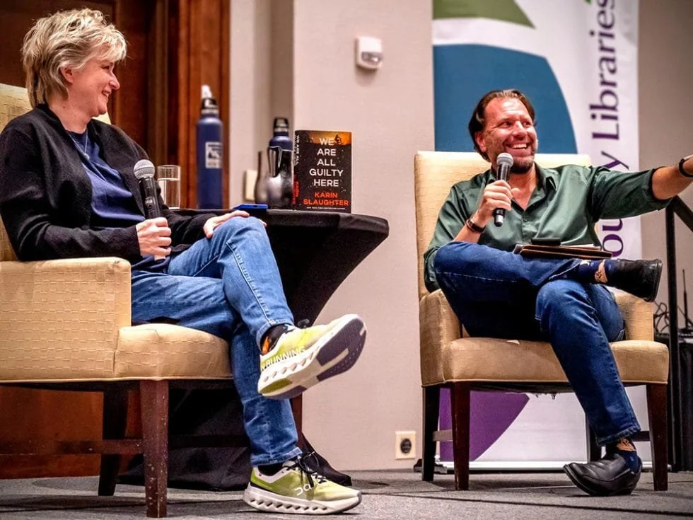 A man and woman sitting in armchairs on a stage each holding a microphones, with a book on the table between them with the title: 