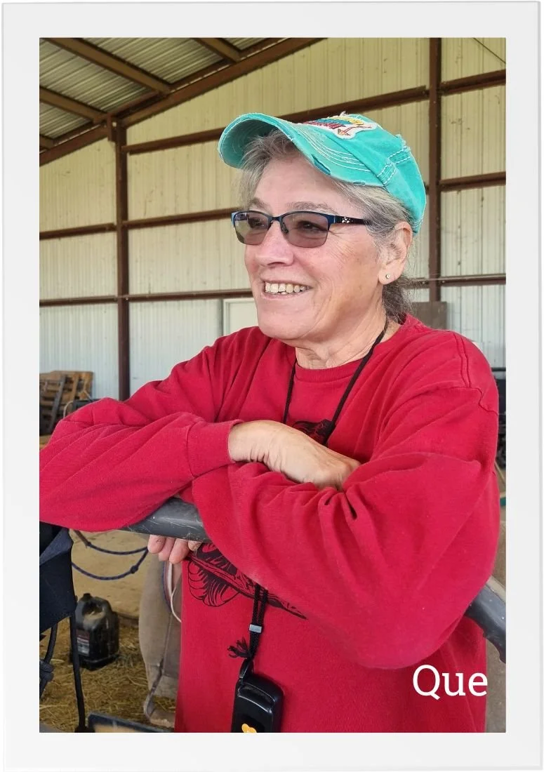 Que, Barn Manager at HorseSpeak, leaning on a fence inside the barn, smiling and relaxed during a quiet moment on the ranch.