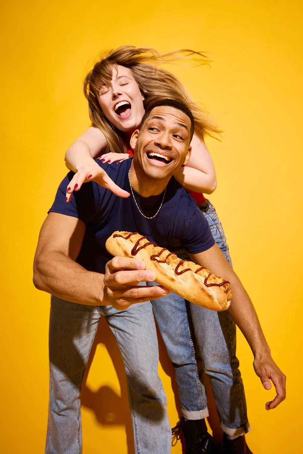 A laughing couple sharing a piggyback and a hotdog in Rollover’s yellow studio campaign, photographed by Jon Enoch.