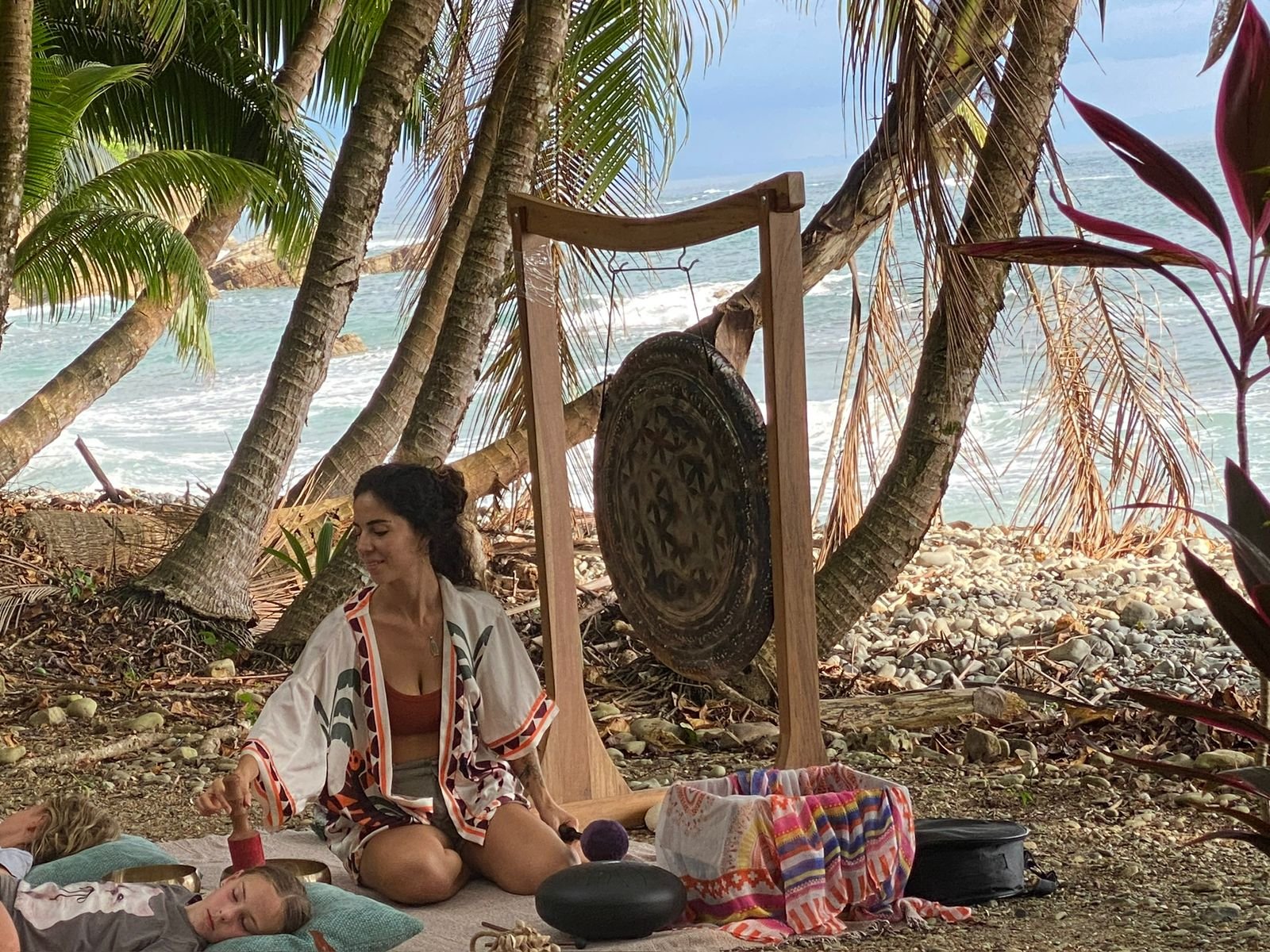 Yoselyn playing singing bowls beside a gong beneath palms by the ocean