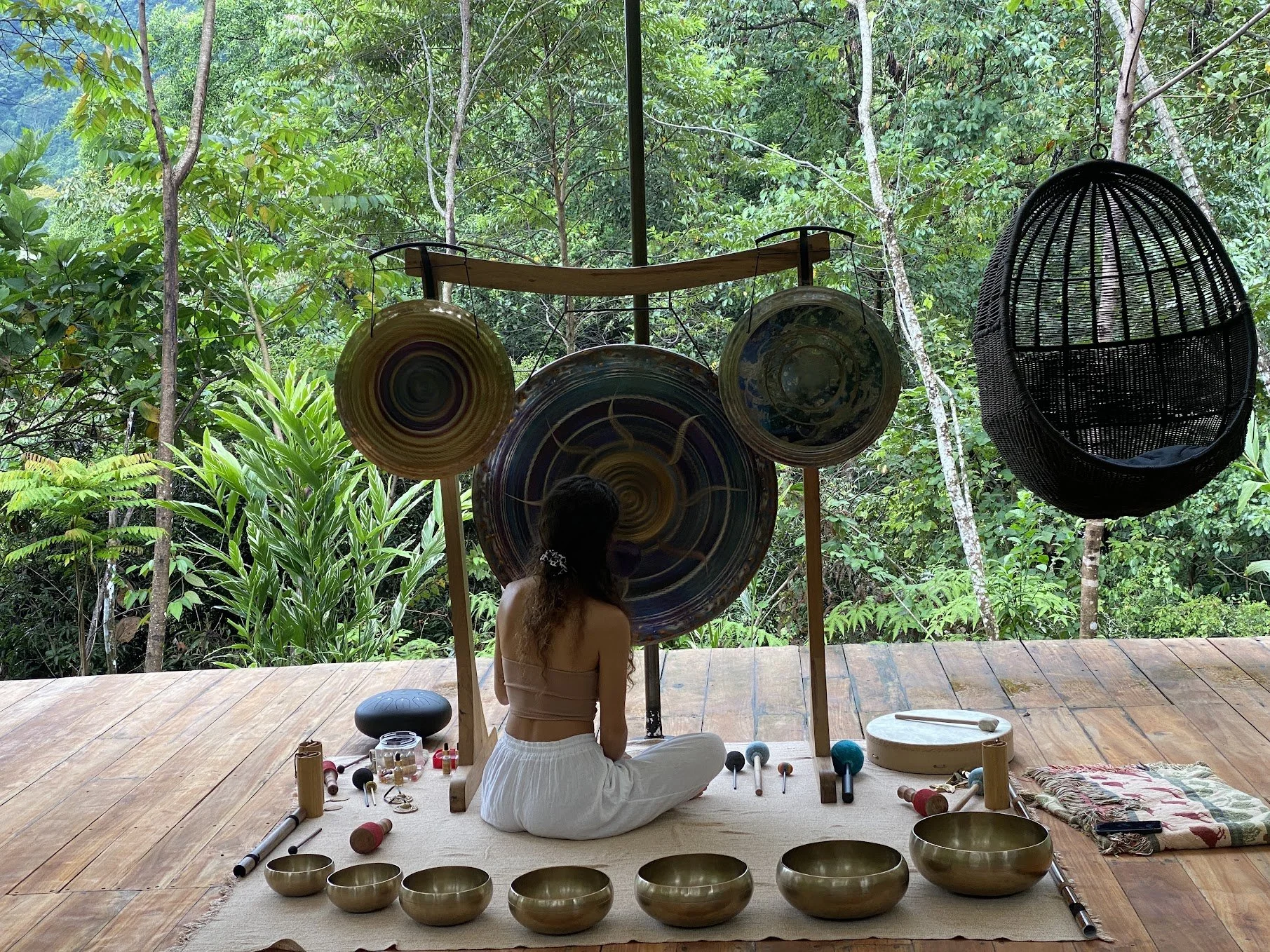 Yoselyn seated with gongs and singing bowls on a forest-facing deck