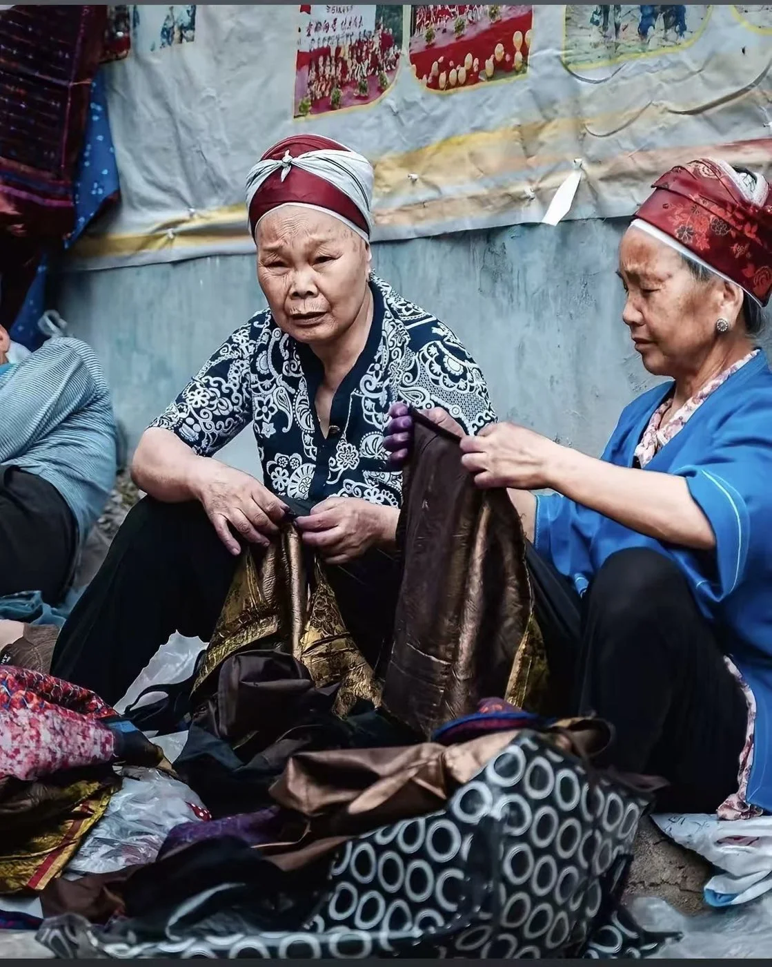 Two elder women in headwraps sorting bundles of patterned and shimmering cloth at a market