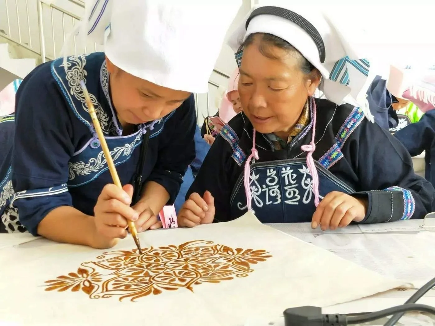 An experienced maker guides a trainee painting a floral resist pattern onto white cloth