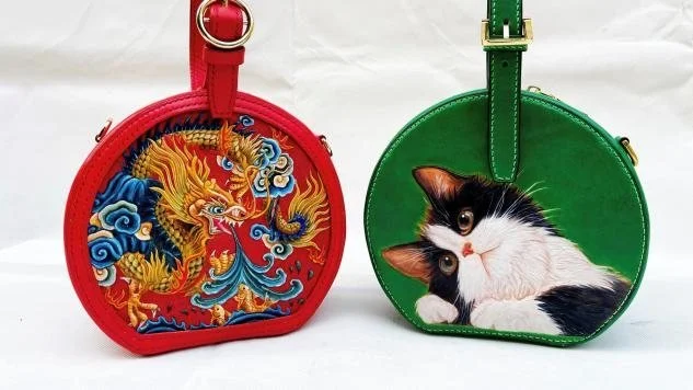 Two round leather bags, one red with a hand-painted dragon and one green with a hand-painted cat