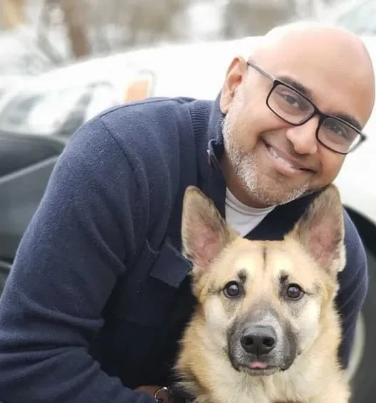 A man with glasses smiling while holding a dog with pointy ears and a tan coat outdoors near Vehicles.
