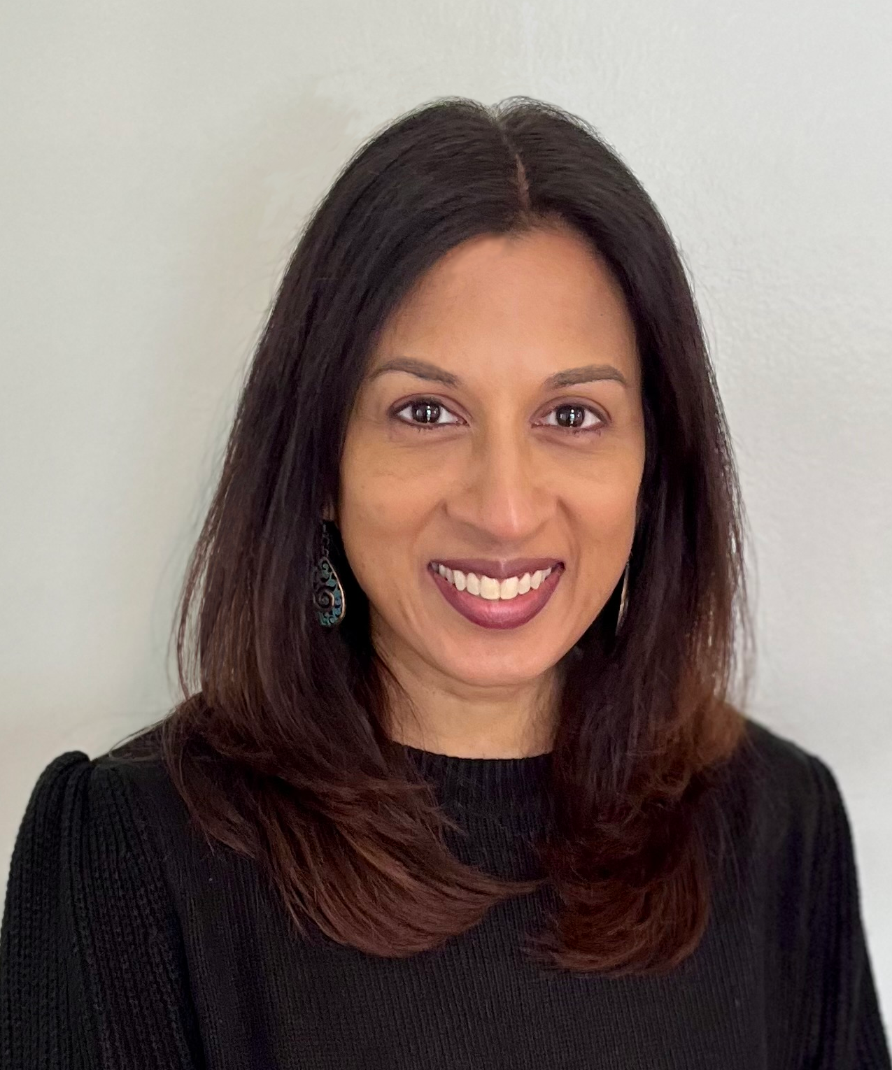 A woman with shoulder-length dark brown hair, wearing a black top and turquoise earrings, smiling against a plain white wall.
