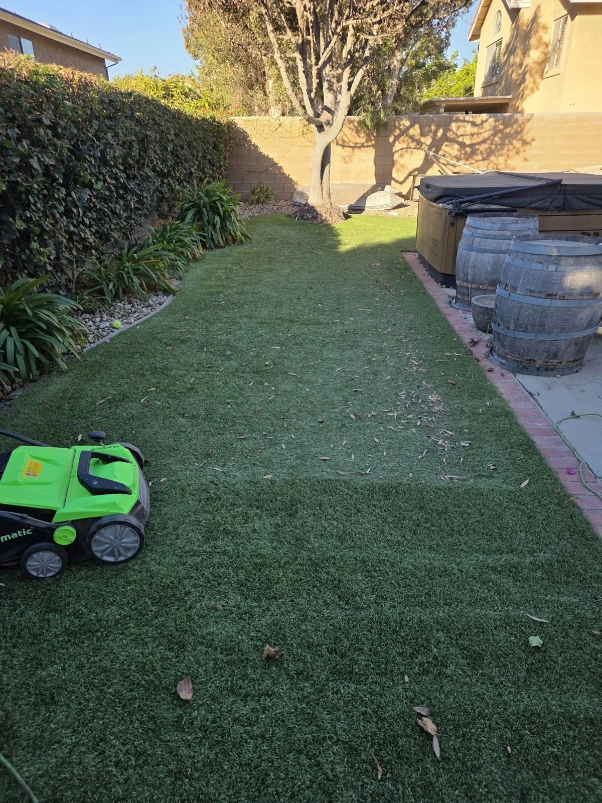 Side-yard artificial turf mid-service: the near half brushed clean, the far half still covered in leaves, with the power brush in frame