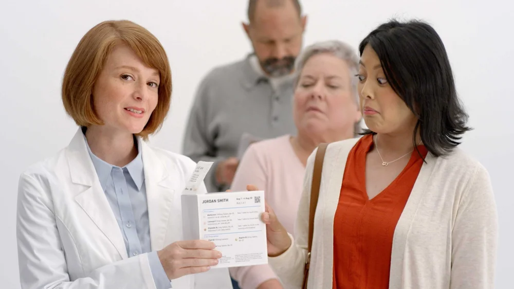 A woman in a white lab coat hands a woman a PillPack box standing in a pharmacy line on a white studio background in PillPack's website explainer video