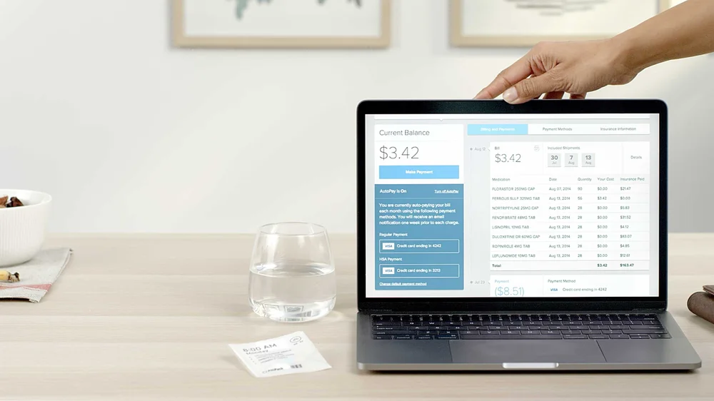 A hand opens a laptop on dining room table showing the PillPack website