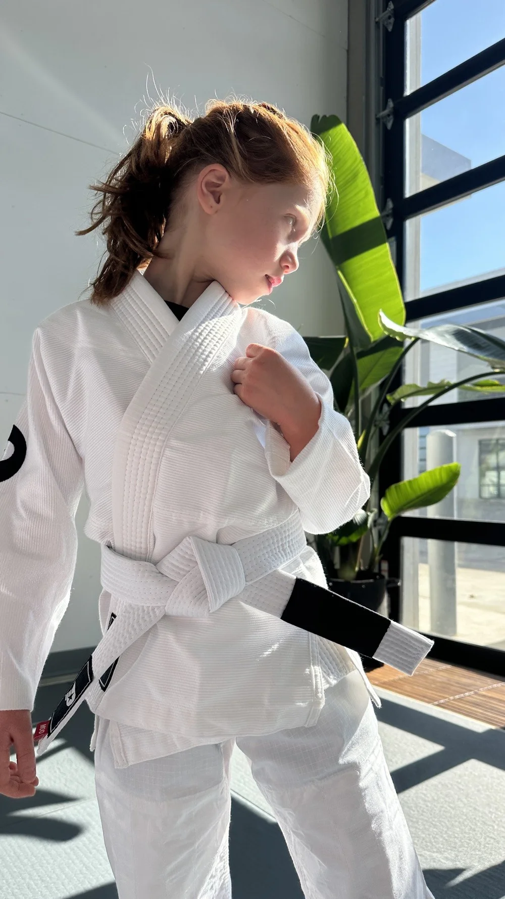 A young girl in a white gi adjusting her lapel in the sunlight at Practice Jiu Jitsu in Georgetown, TX