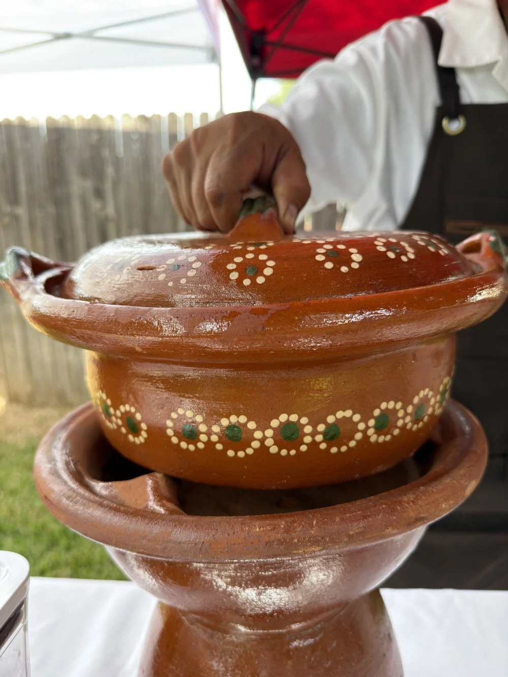 A hand lifting the lid off a painted clay pot of meat at a Mesa y Maiz spread