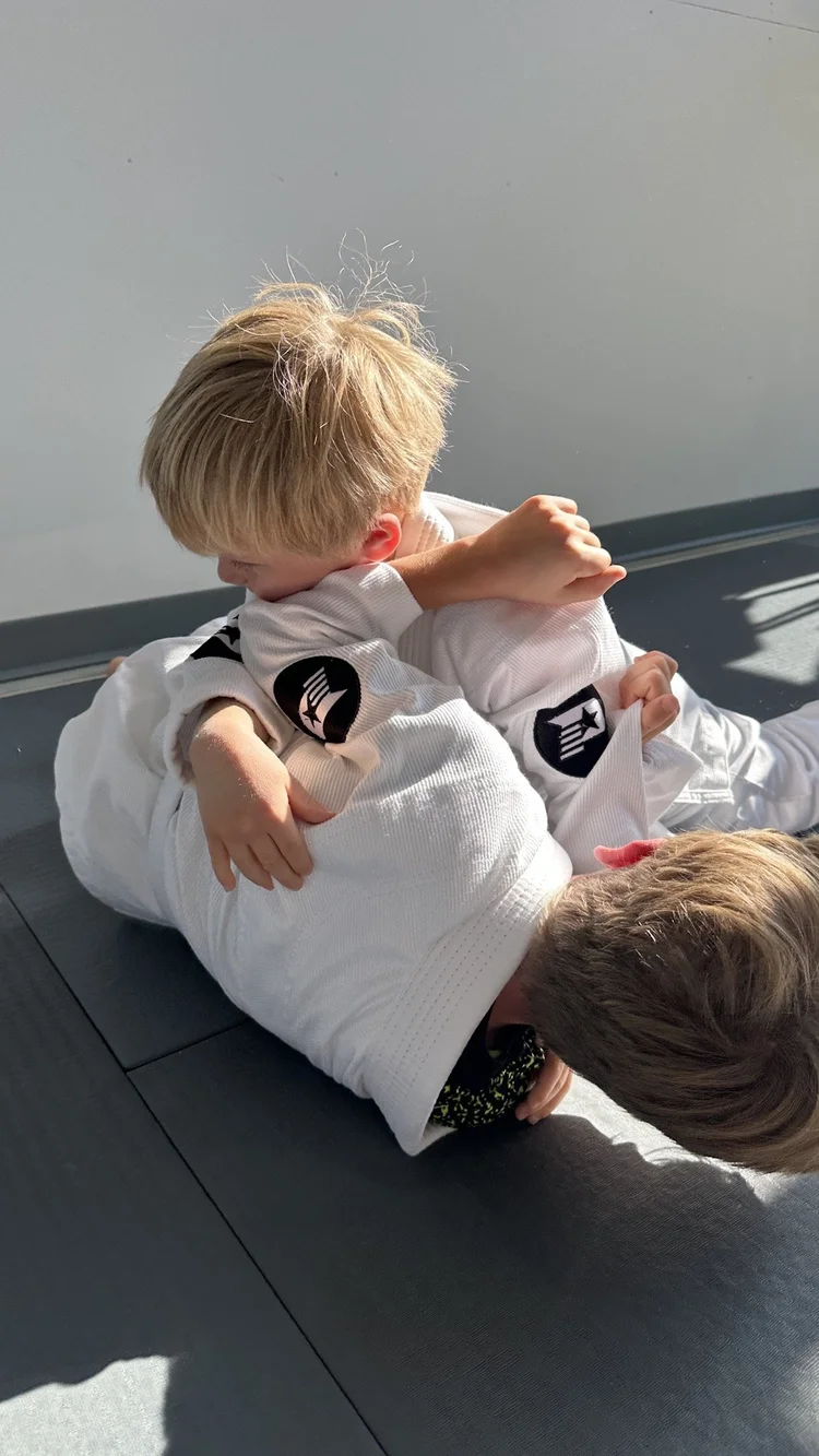 Two preschool-age children in white gis playing and rolling together on the mat at Practice Jiu Jitsu in Georgetown, TX