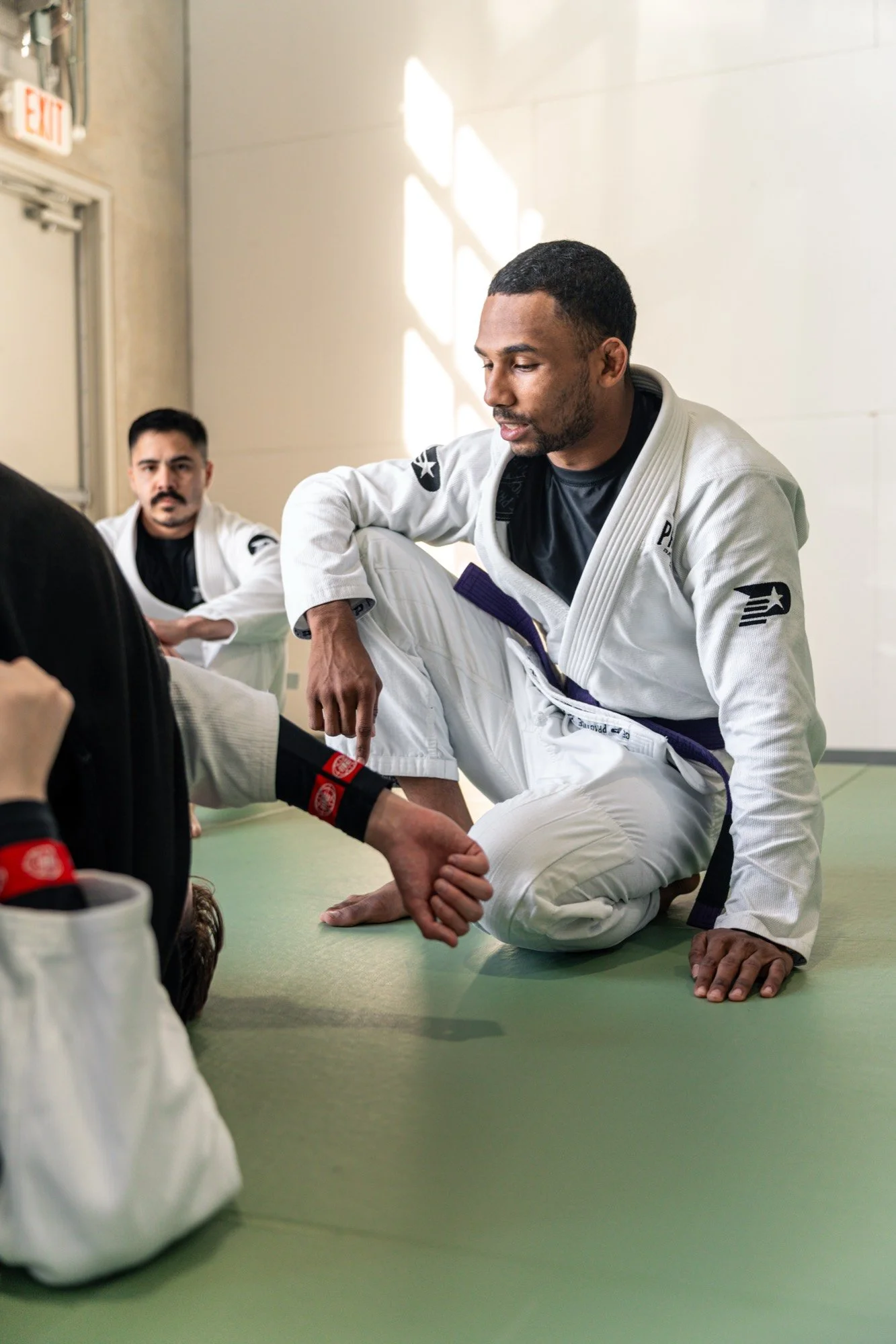 An adult student in a Practice Jiu Jitsu gi sitting on the mats in the window light during a class in Georgetown, TX