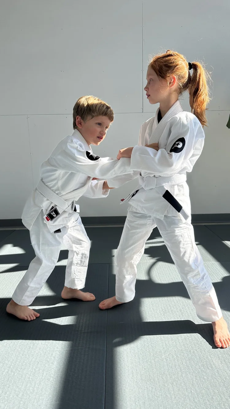Two young children in white gis practising a standing grip drill together at Practice Jiu Jitsu in Georgetown, TX