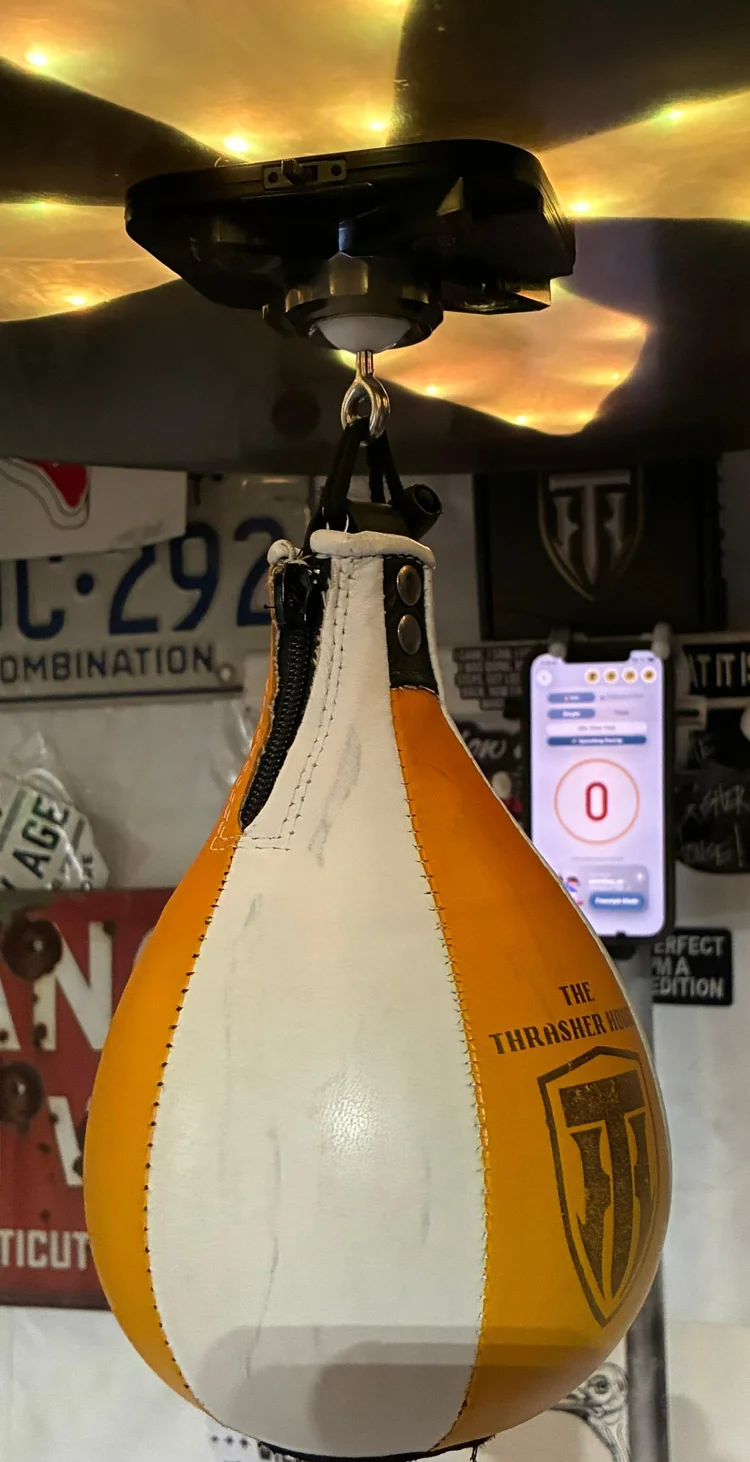 A yellow and white Thrasher speed bag under the counter in a garage hung with license plates, the app open on a phone