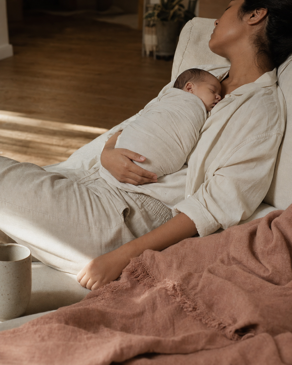 A new parent reclined on a cream linen sofa, a swaddled newborn asleep on their chest, a dusty rose throw spilling across the lower frame.