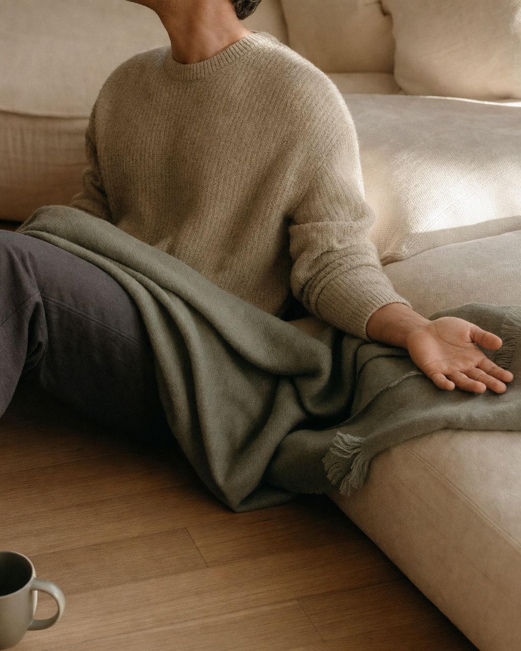 A partner seated on the floor beside a cream linen sofa, leaning back gently, one open hand resting on the sofa cushion, a sage-green throw draped across their lap.