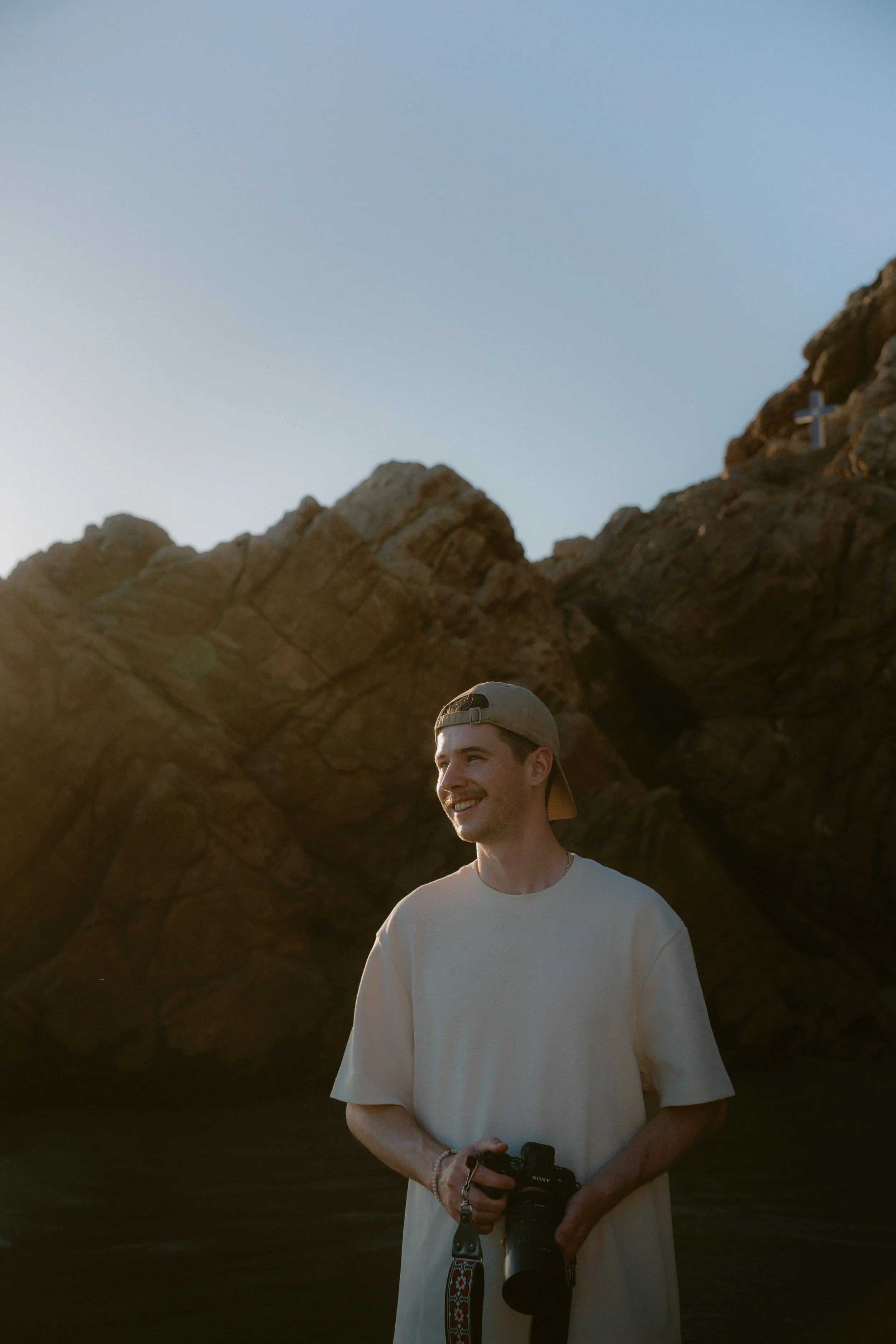 Cam smiling while holding his camera in front of sunlit rocks