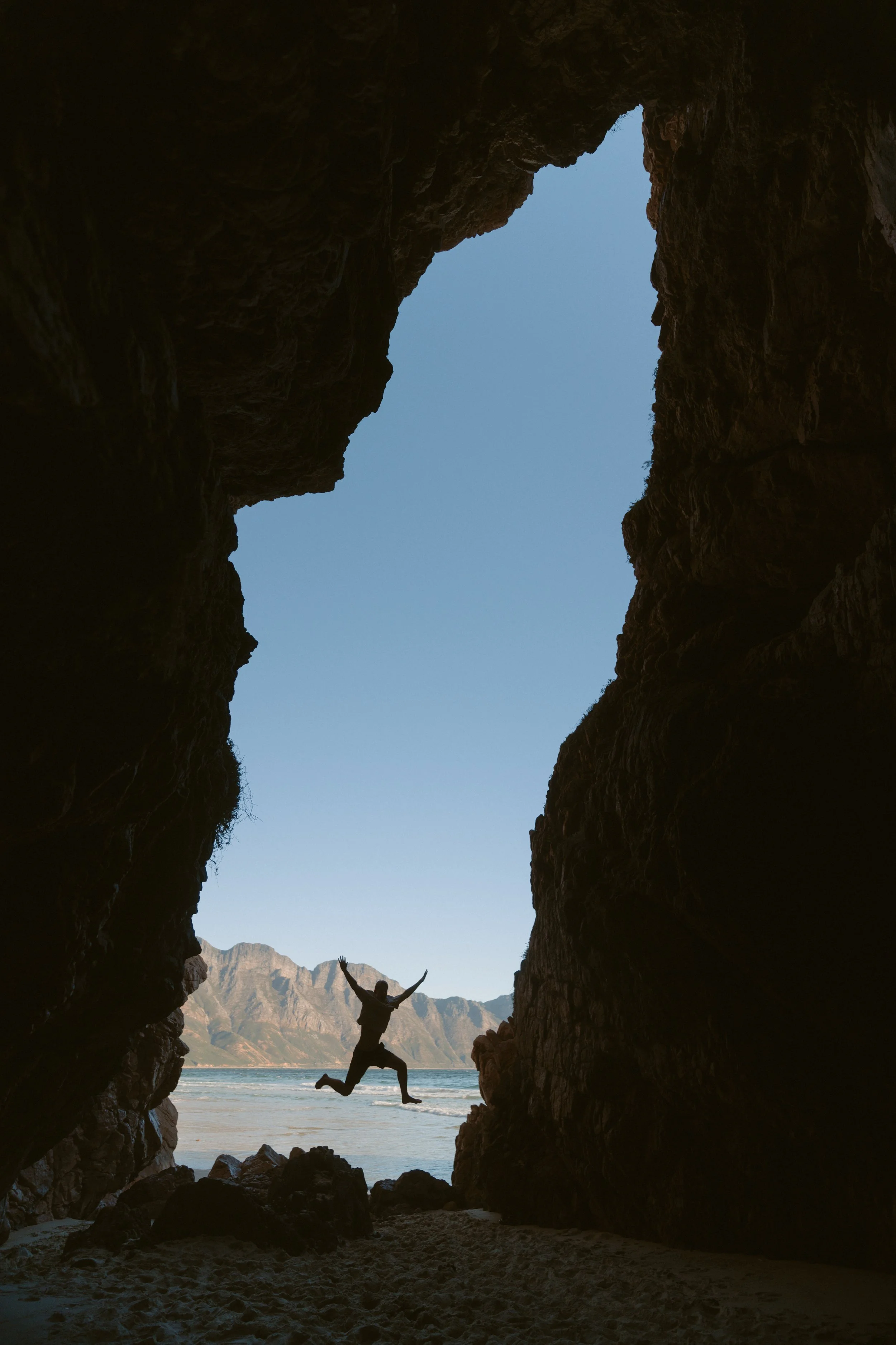 Cam jumping in the opening of a sea cave, with water and mountains beyond