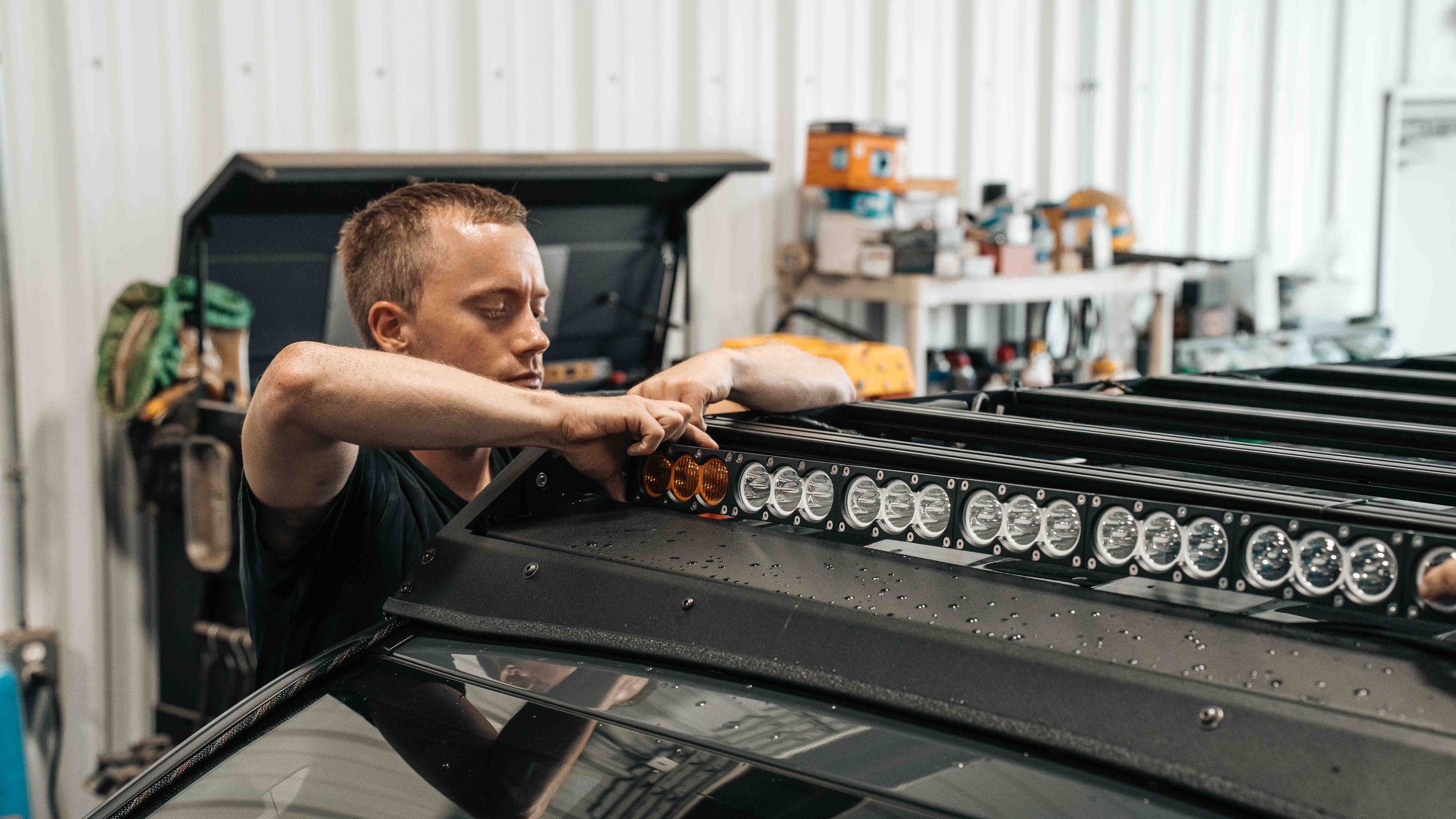 Installer positioning an LED light bar into the front fairing of a full-length roof rack mounted on a vehicle roof.