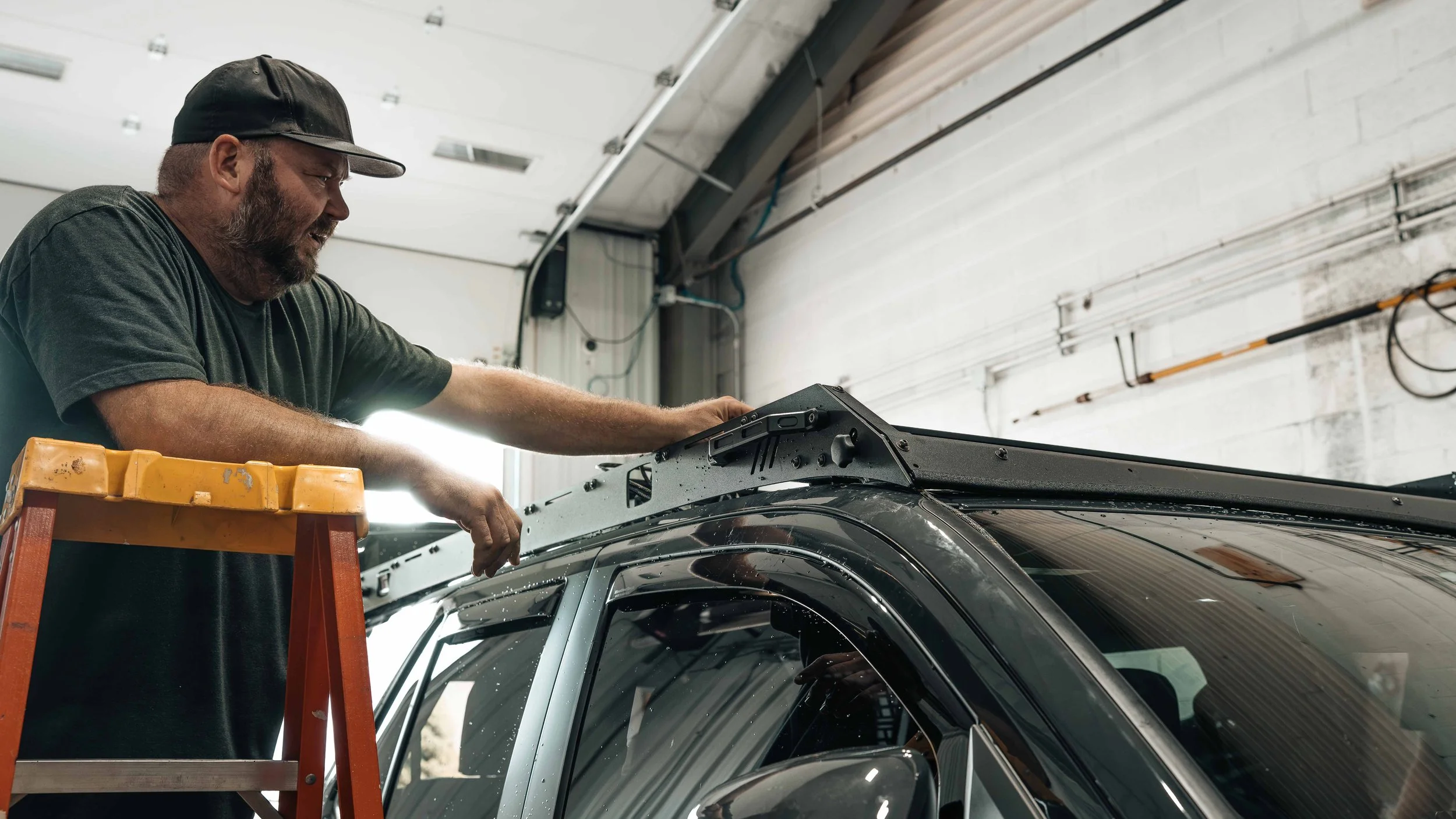 Xtreme technician on a step ladder fitting a low-profile platform roof rack to the roof of a dark SUV inside the Bridgeville, PA install bay.