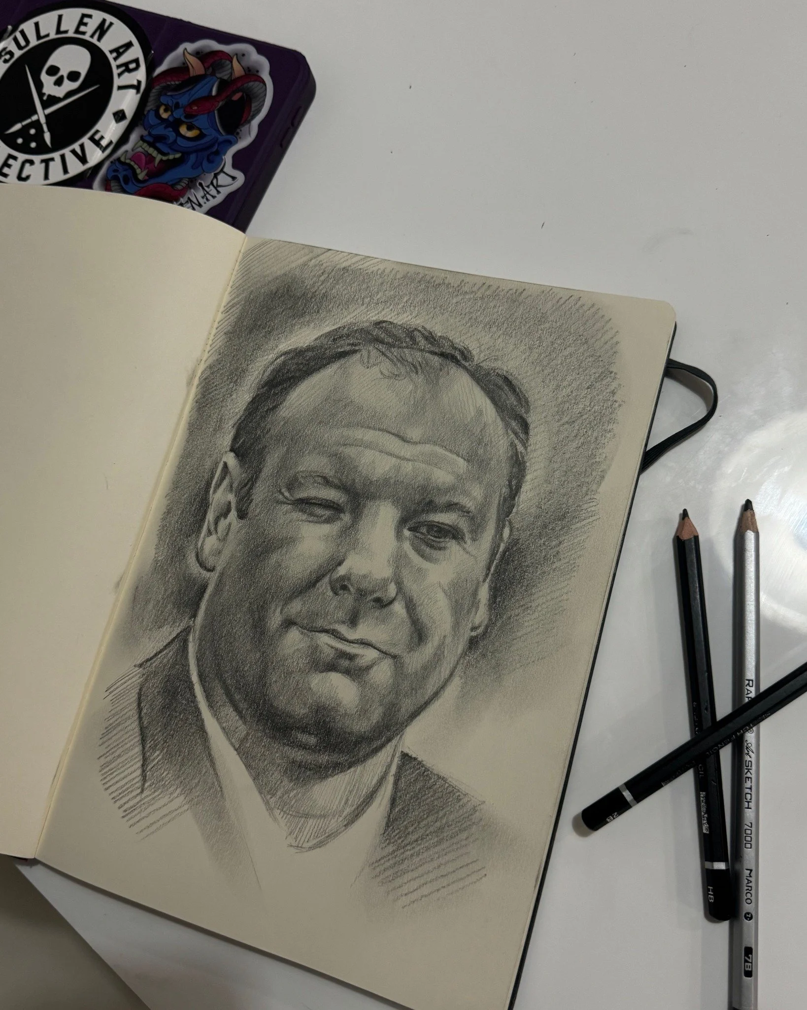 Pencil portrait of a man in a sketchbook, with drawing pencils beside the page.