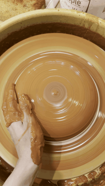 Jael's hands shaping wet red clay on the pottery wheel