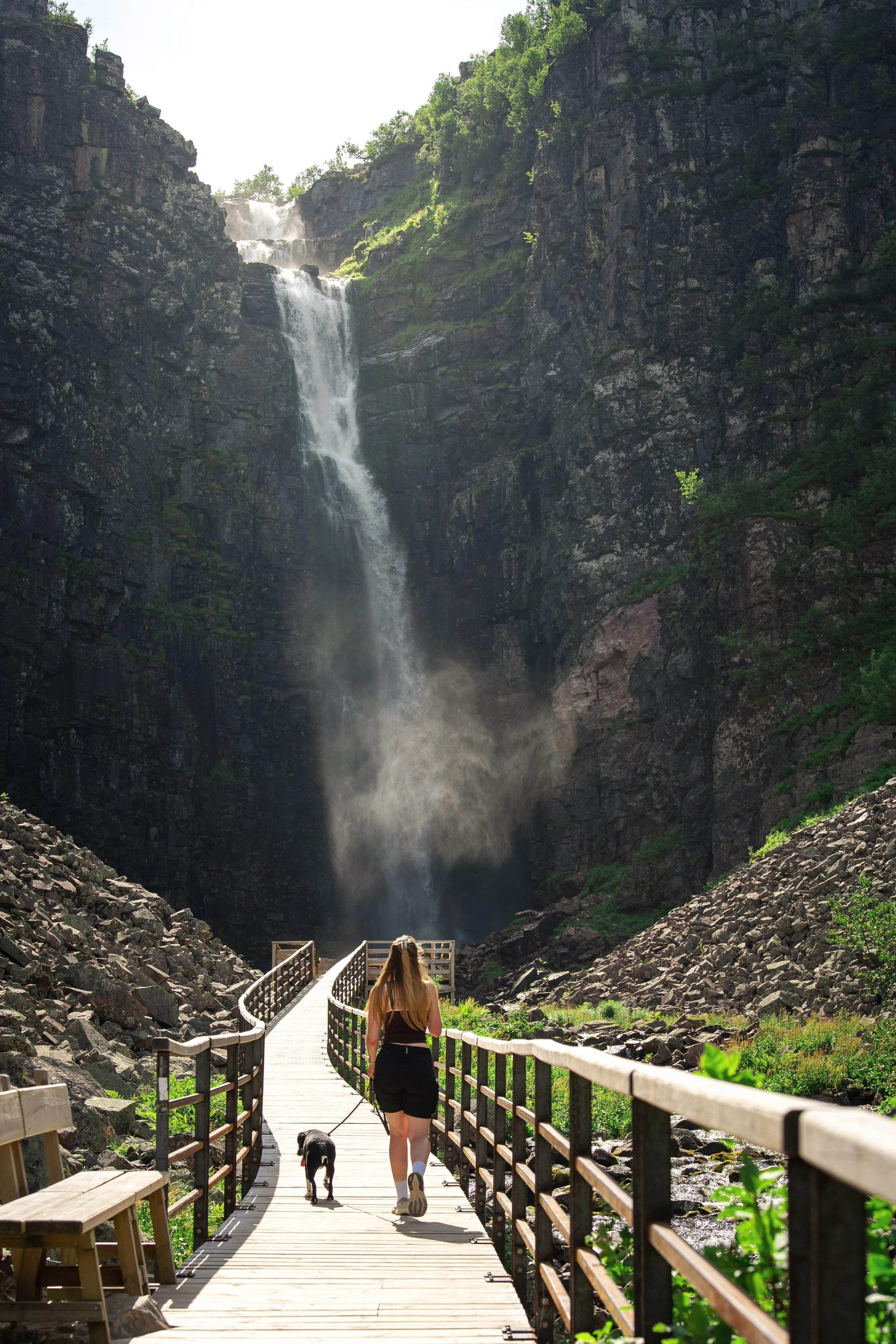 Frau und Hund auf einem Holzsteg vor einem Wasserfall