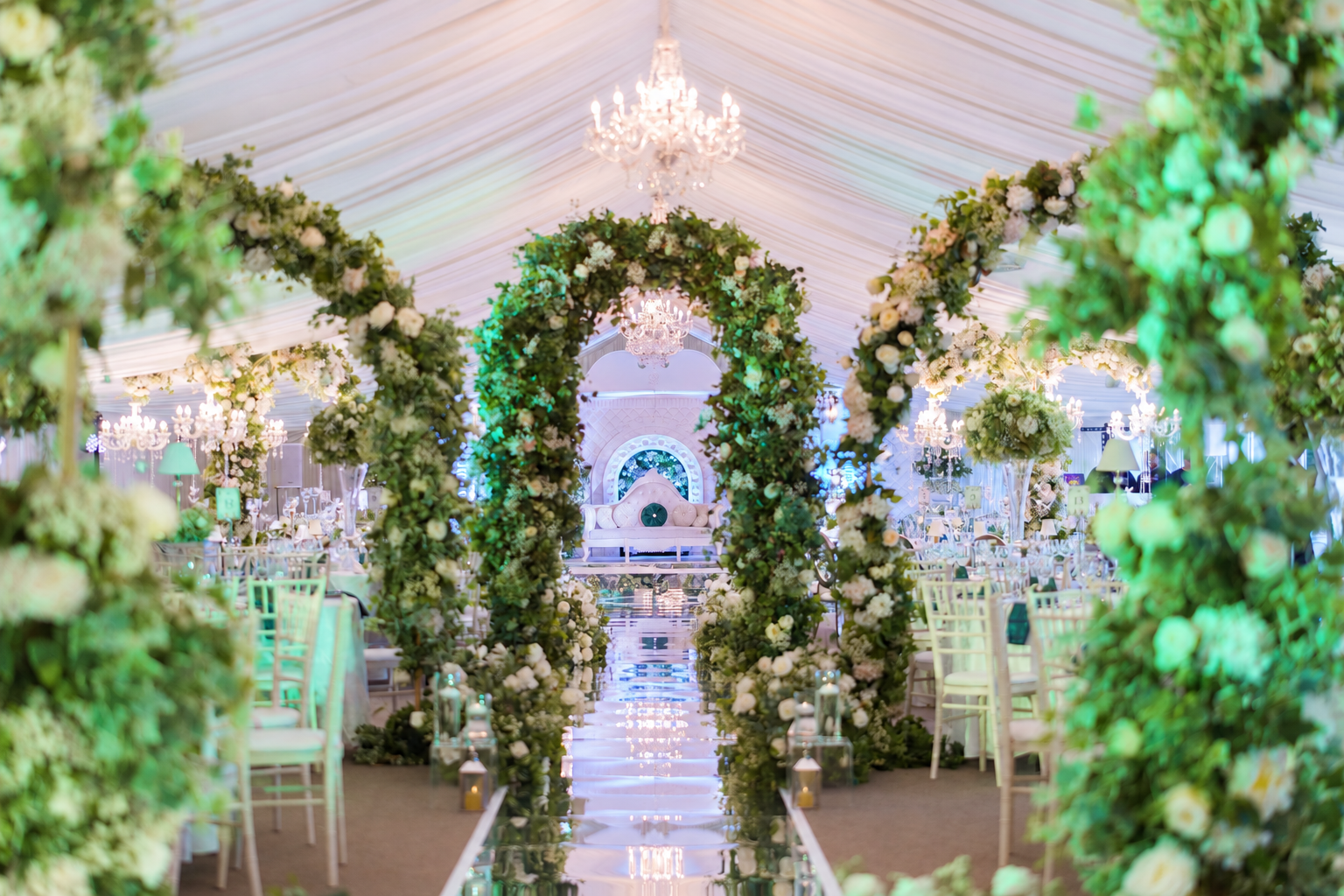 Green and white floral arches framing a wedding aisle beneath a chandelier-filled marquee