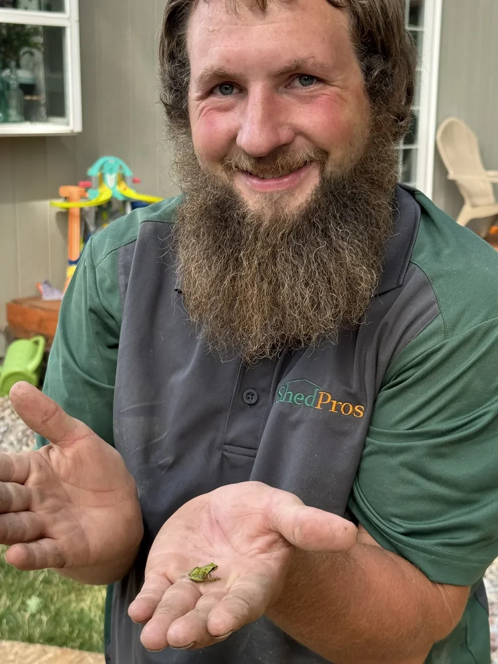 A smiling ShedPro in a company shirt holding a small green frog on a residential job site