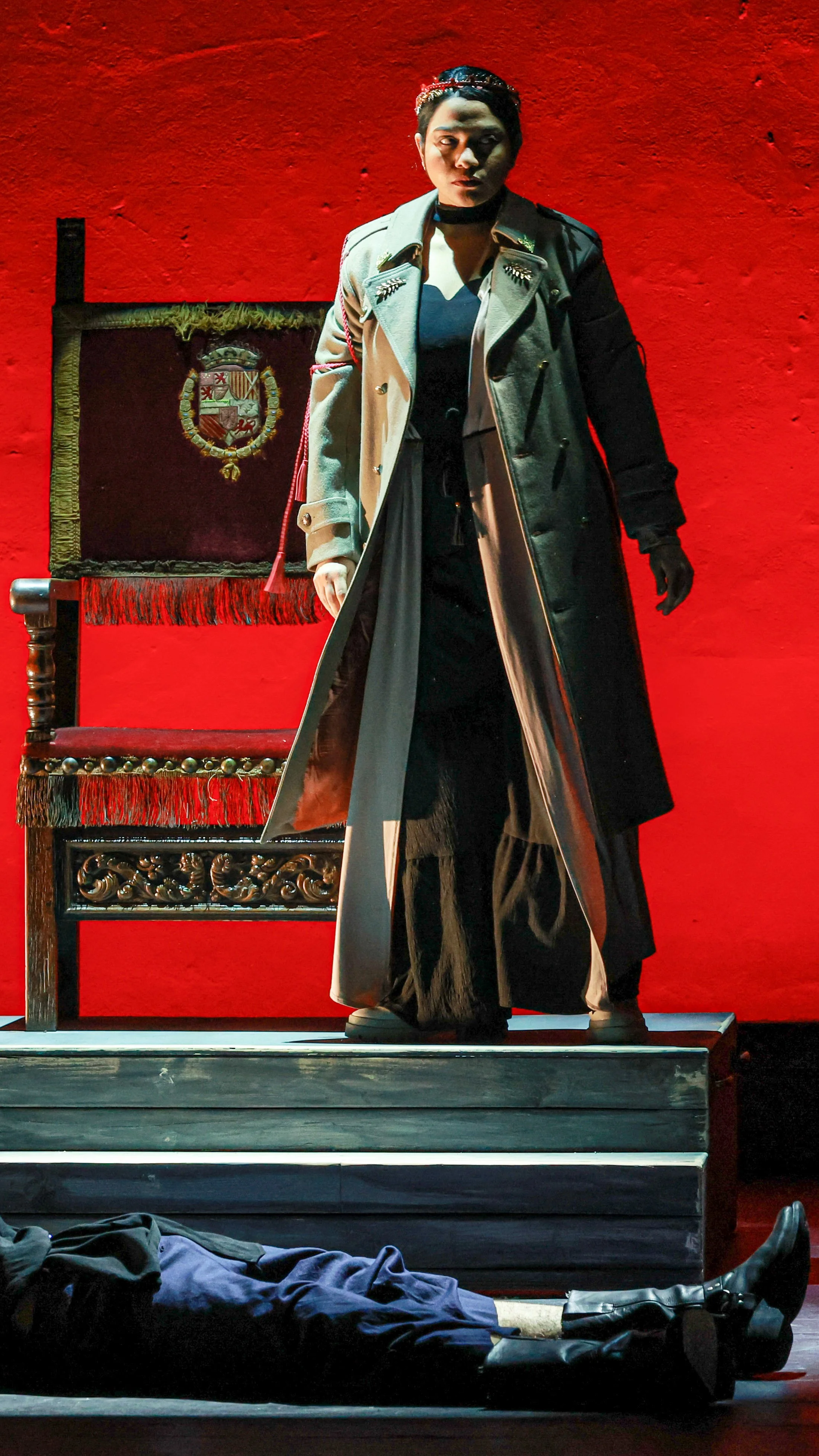 Performer in a long black coat stands before a red throne as another lies onstage in LEAR.