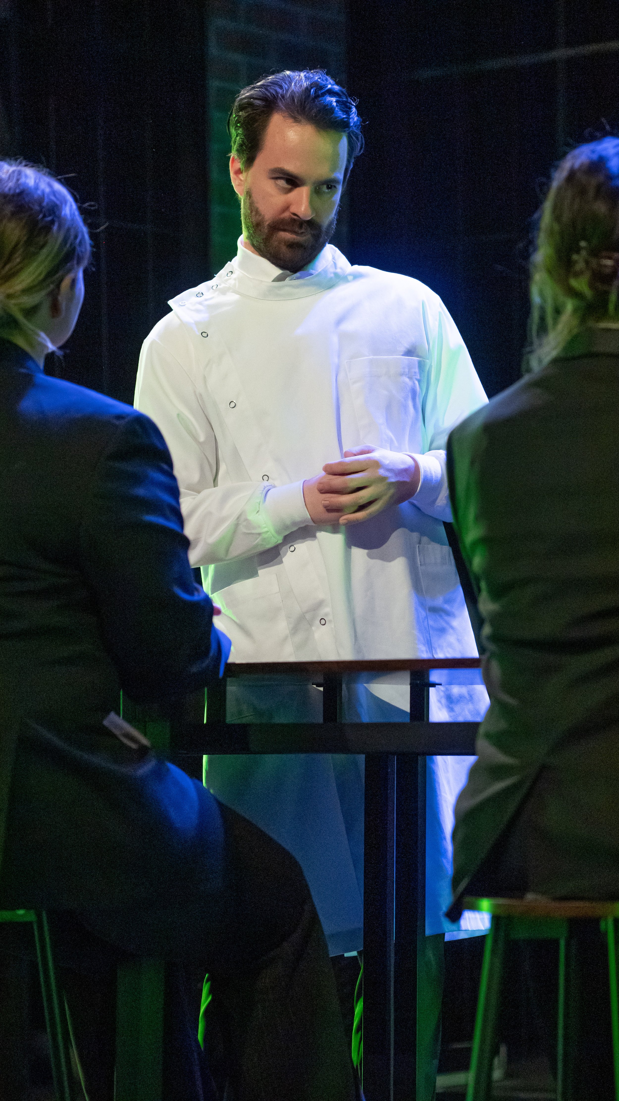 Performer in a white coat stands before two seated figures in Radium Girls.