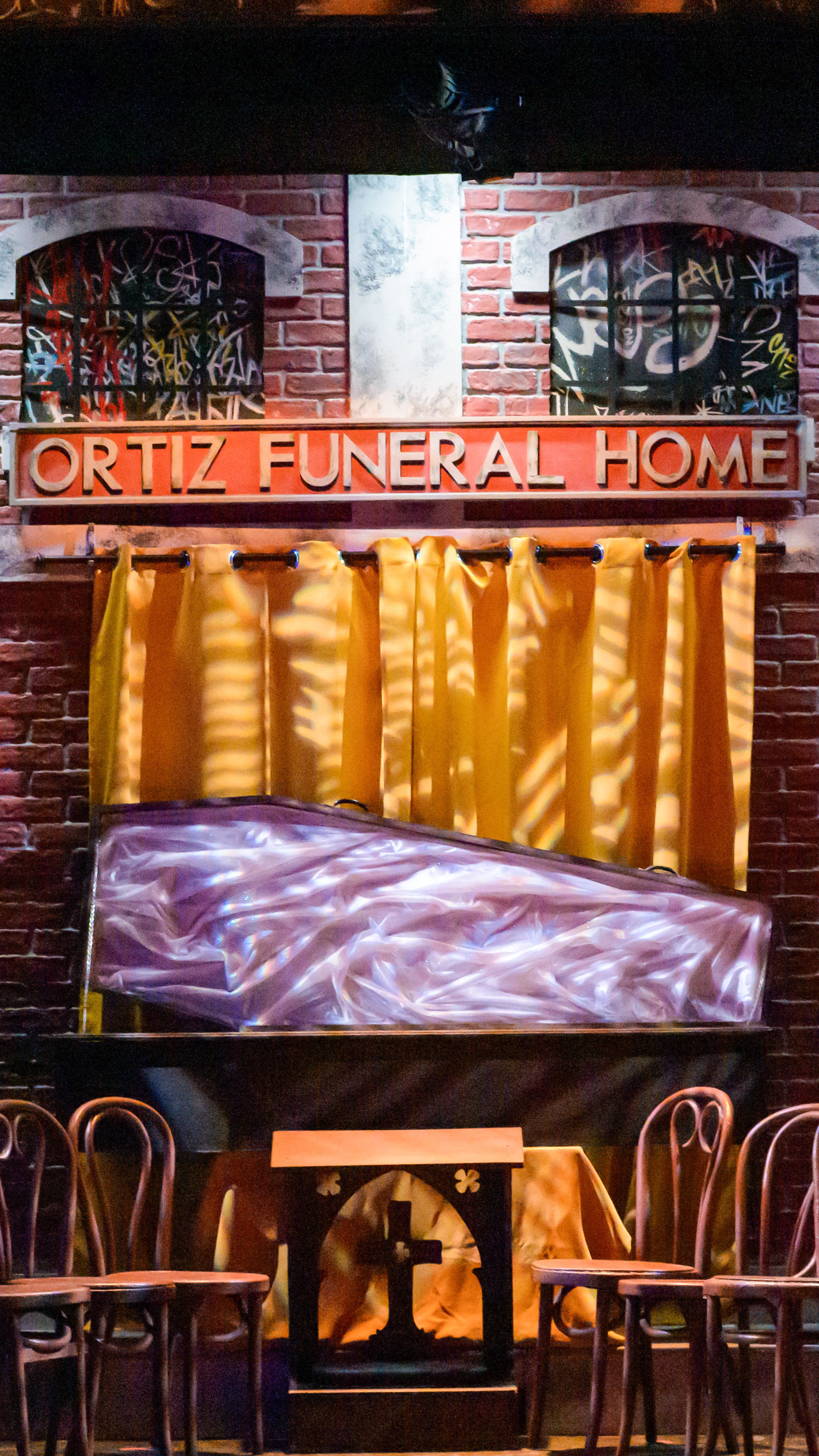 Funeral home set with brick walls, gold curtains, chairs, and a coffin in Our Lady of 121st Street.