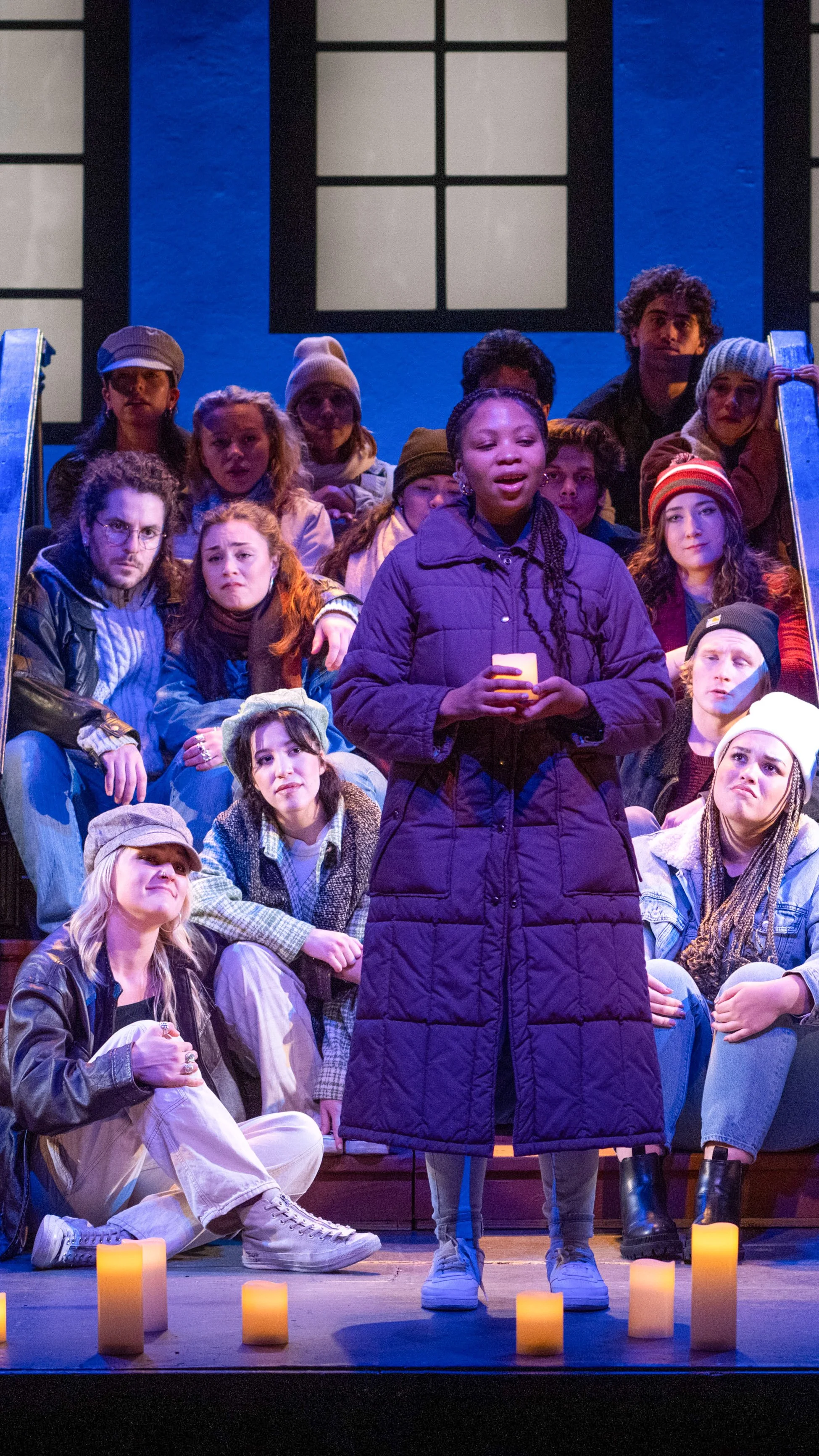 Large ensemble gathers around a performer in a dark coat beside glowing candles in Stoop Songs.