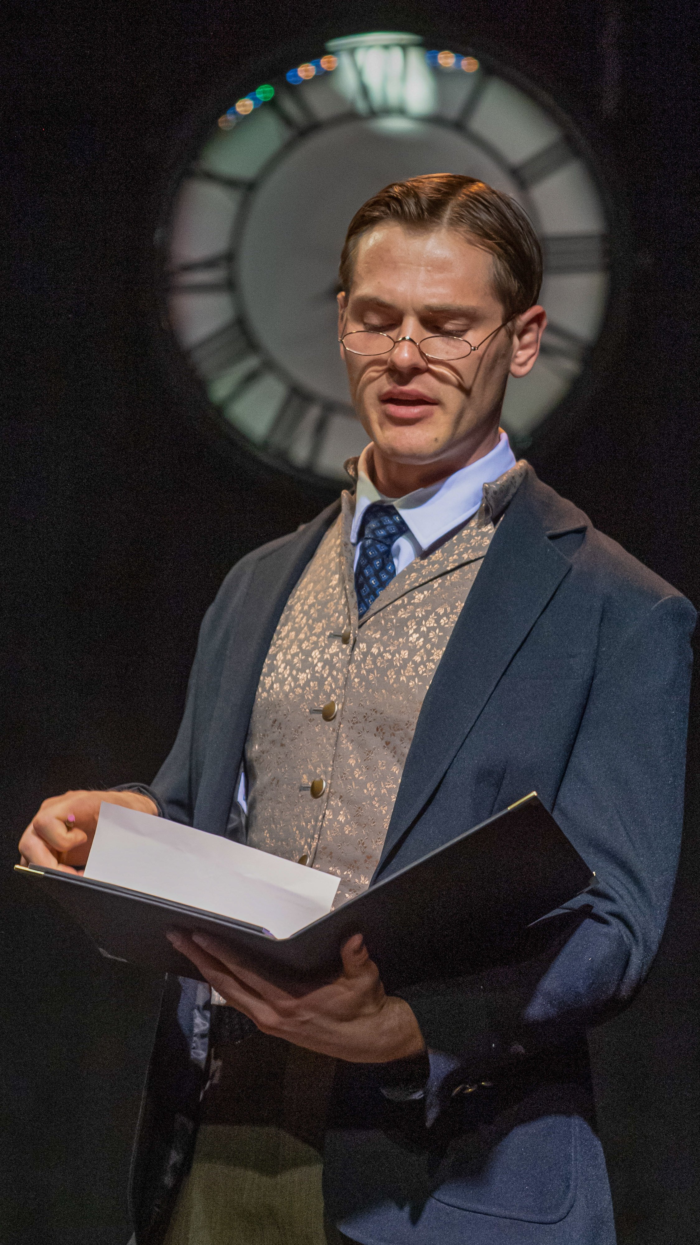 Performer in a suit reads from an open book beneath a large clock in Radium Girls.