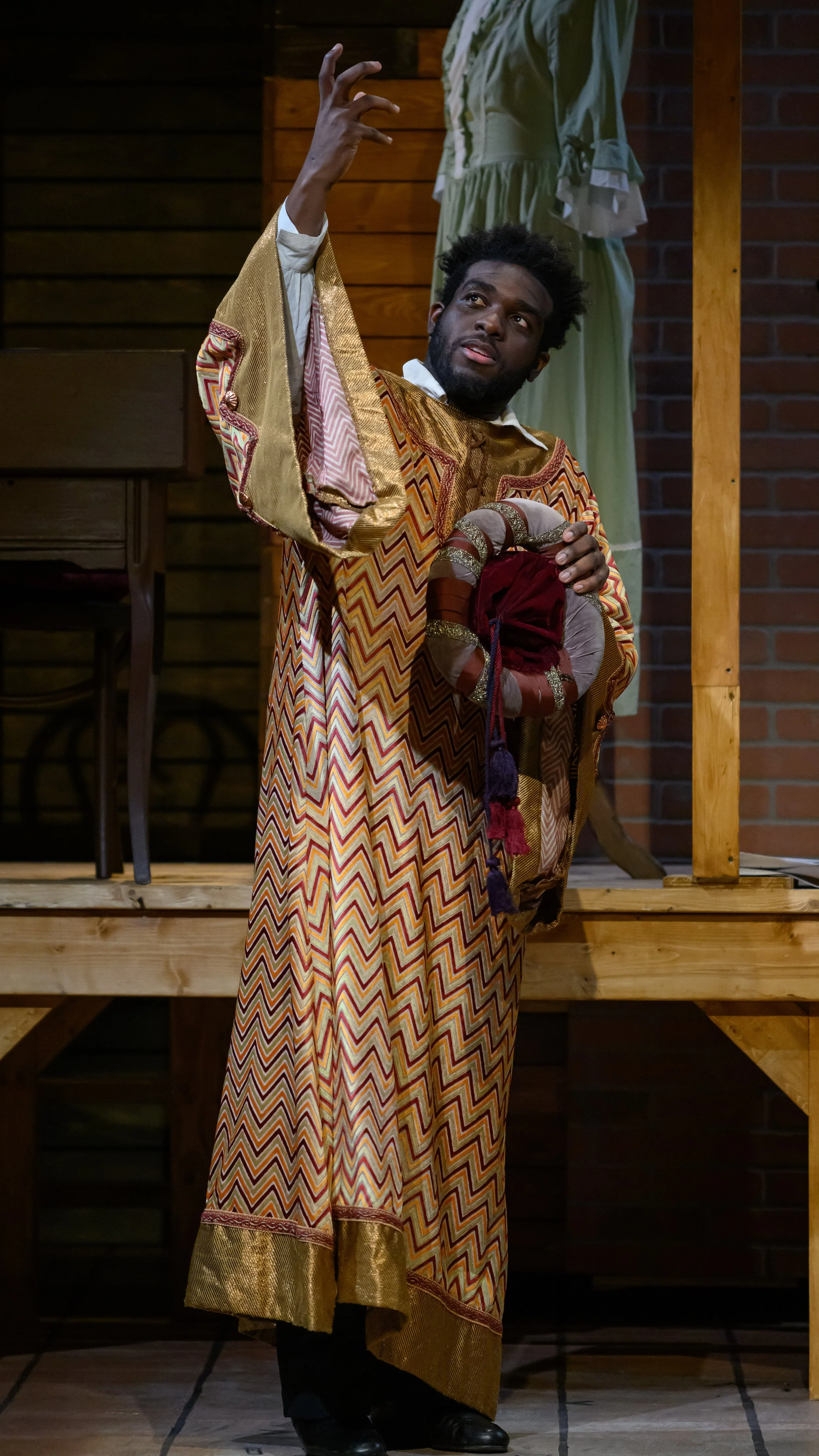 Performer in a patterned robe raises one hand while holding costume fabric in Red Velvet.