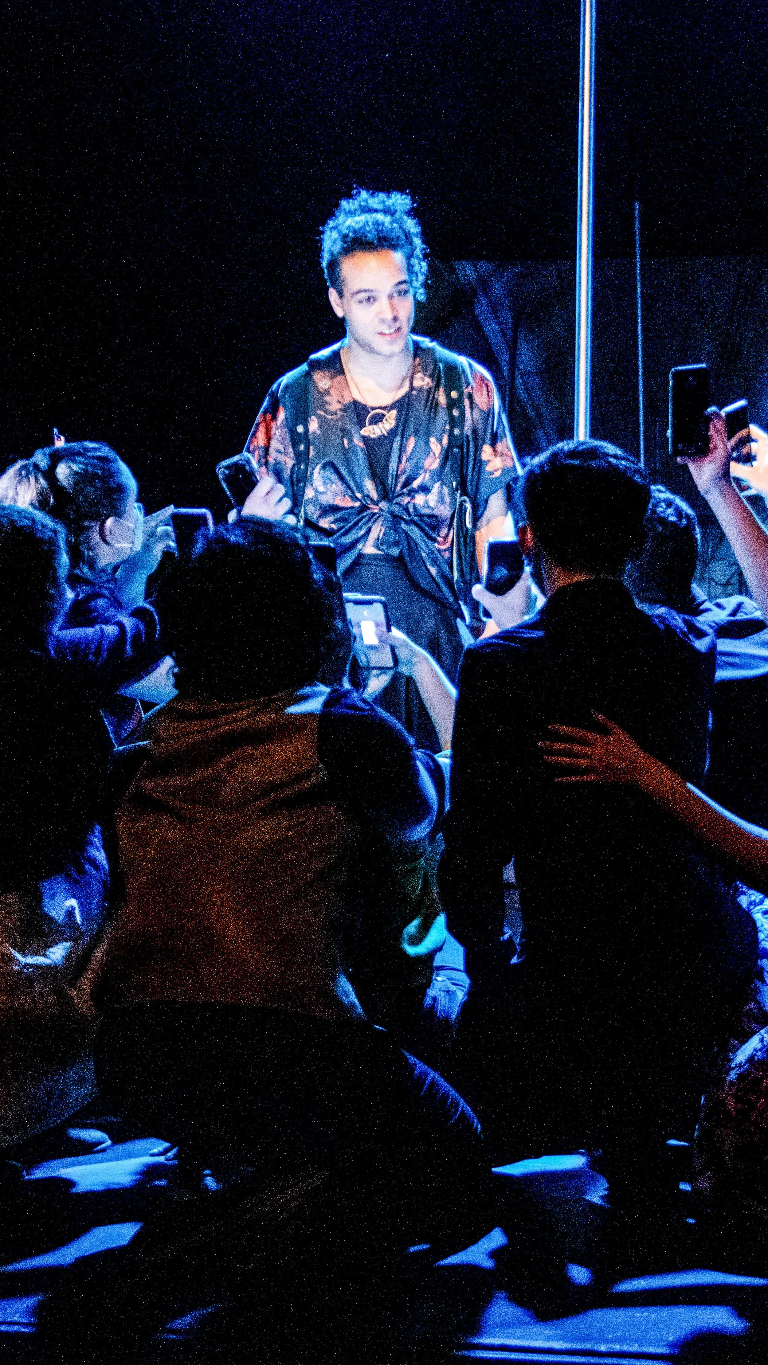Performer in a dark costume stands before a seated ensemble under blue light in Another Hundred People.